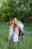 A family gathers together in a lush green outdoor setting, with grass underfoot and leafy trees in the background. The group consists of two adults and four children. The children are dressed in light summer clothing, and one child is being held by an adult.