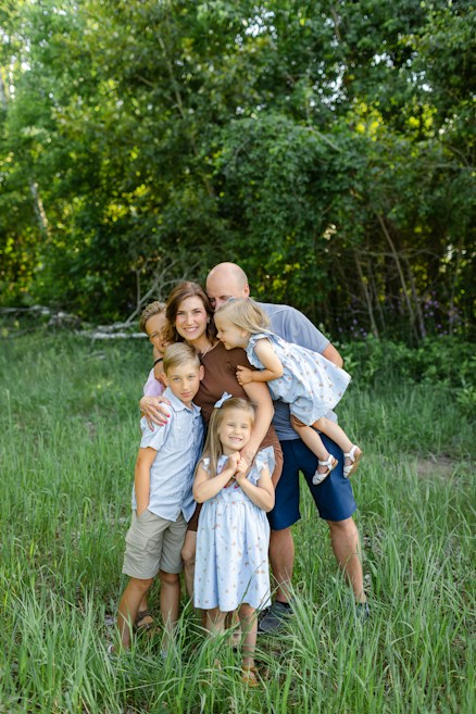 A family gathers together in a lush green outdoor setting, with grass underfoot and leafy trees in the background. The group consists of two adults and four children. The children are dressed in light summer clothing, and one child is being held by an adult.
