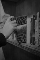 A hand is reaching for a book on a wooden bookshelf. The book's cover displays the name 'Susan Hill', and it appears to be a sequel related to 'Rebecca.' Other books are also visible on the shelf, with different titles and colors, but are mostly obscured. The setting is cozy and suggests a library or a personal study.
