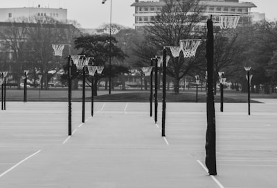 A series of empty outdoor basketball courts with numerous hoops aligned symmetrically. The background features bare trees and city buildings, creating a stark and somewhat desolate atmosphere.