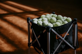 A large black tennis ball hopper filled with numerous yellow-green tennis balls is standing on a clay tennis court. The floor of the court has a warm, reddish-brown color, with soft, angular shadows falling across it, suggesting sunlight streaming in from above.