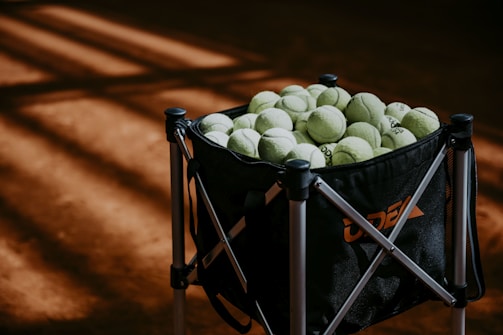 A large black tennis ball hopper filled with numerous yellow-green tennis balls is standing on a clay tennis court. The floor of the court has a warm, reddish-brown color, with soft, angular shadows falling across it, suggesting sunlight streaming in from above.