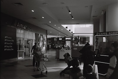 A black and white photograph of a shopping mall interior. The image includes a life clinic on the left side, with signage for various clinics like pharmacy and diabetes clinic. A person is pushing a shopping trolley, and several people are visible walking around, including someone with a stroller. Shops and a cafe kiosk can be seen in the background.