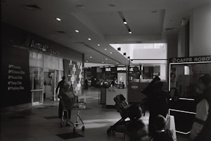 A black and white photograph of a shopping mall interior. The image includes a life clinic on the left side, with signage for various clinics like pharmacy and diabetes clinic. A person is pushing a shopping trolley, and several people are visible walking around, including someone with a stroller. Shops and a cafe kiosk can be seen in the background.