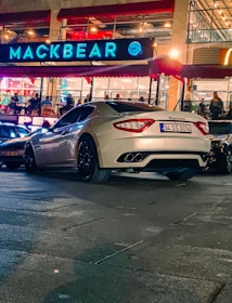 A luxury white sports car is parked on a city street. The car is positioned near a well-lit restaurant with a large sign reading 'MACKBEAR'. Neon lights and reflections illuminate the scene, enhancing the urban nighttime atmosphere.