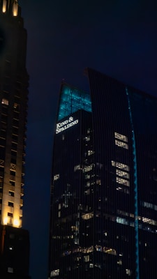 Two tall buildings in an urban area are seen at nighttime. One building has a prominent sign that reads 'King & Spalding', with bright lights illuminating a section of the building. The other building is partially visible and has small lights scattered across its exterior. Both structures stand against a dark, night sky.