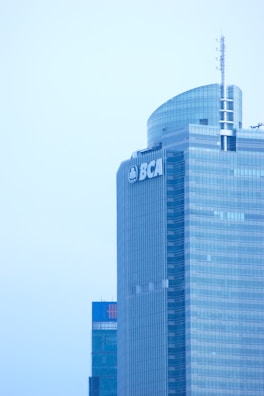 A tall, modern skyscraper with reflective glass windows and a rounded top structure. The building features a prominent logo with the letters BCA near the top. In the background, there's another high-rise building with a logo visible near the roofline.