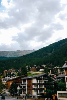 A mountain village with multiple chalet-style buildings surrounded by lush green forests. The buildings have wooden facades and are built on various levels on the hillside. Above, there is a cloudy sky with patches of light breaking through.