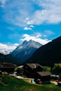 A picturesque mountain landscape with a clear blue sky and fluffy clouds. A village with wooden houses is located in the foreground, surrounded by lush green hills and a majestic snow-capped mountain rising in the background.