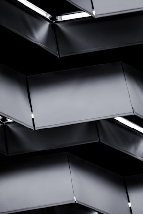 A series of geometric, metallic panels with a sleek, modern design. The panels are layered, creating a repeating pattern with highlights of light reflecting off their surfaces. The structure has a clean, minimalistic aesthetic.