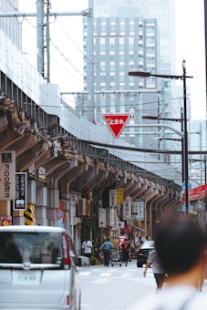 An urban street scene in Japan with buildings in the background, elevated train tracks, and various signs in Japanese. The street is bustling with cars and pedestrians, and there is a prominent red yield sign.