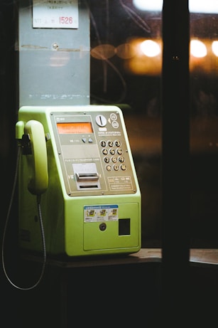 A green public telephone is illuminated in a dimly lit area, showcasing its keypad, coin slot, and handset. The background is slightly blurred, with warm lights in the distance creating a cozy ambiance.