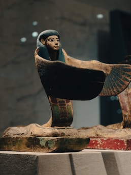 An ancient Egyptian artifact depicting a figure with a human face and large wings. The figure appears to be a statue or carving made from stone or clay, with faded colors suggesting age. It is positioned on a pedestal, possibly in a museum setting.