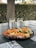 A platter filled with various snacks including slices of salami, cheese cubes, cucumber slices, and cherry tomatoes. Doritos chips are scattered around the platter. Two glasses of water with lemon slices are placed behind the platter on a dark table. The setting appears to be outdoors with a background of a tree trunk and dense greenery.