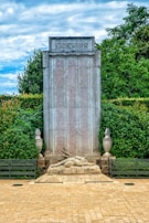 A large stone monument stands tall in front of a lush green hedge. The monument features a list of names etched in vertical columns. At the top of the monument, there is an engraving with dates and text above the names. At the base, a stone statue of a reclining soldier is depicted, with some additional text inscribed. The area is paved with light-colored bricks, and the adjacent environment appears serene and well-maintained.