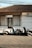 Two motor scooters are parked in front of a storefront with a tiled roof. The building has a closed sliding shutter on one side and a glass door with curtains on the other, above which is a sign that reads 'SHERLY Shop'. The pavement is patterned, and there are power lines visible overhead.