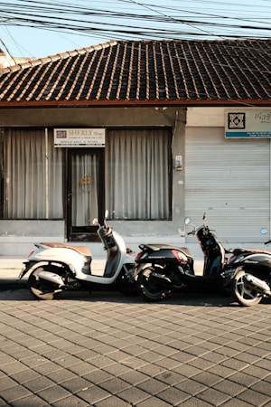 Two motor scooters are parked in front of a storefront with a tiled roof. The building has a closed sliding shutter on one side and a glass door with curtains on the other, above which is a sign that reads 'SHERLY Shop'. The pavement is patterned, and there are power lines visible overhead.