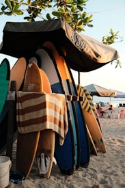 A collection of surfboards rests under a canopy on a sandy beach. Some boards are covered with a striped towel, and the scene is shaded by leafy branches from a nearby tree. In the background, white plastic chairs and tables are visible, positioned to overlook the ocean.