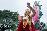 A large colorful statue of the Hindu deity Ganesha, adorned with intricate jewelry and clothing, is carried on a decorated platform. The platform features a large ornate frame with a pink center. Behind the statue, there are lush green trees under a partially cloudy sky.