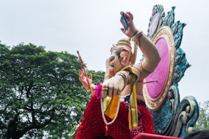 A large colorful statue of the Hindu deity Ganesha, adorned with intricate jewelry and clothing, is carried on a decorated platform. The platform features a large ornate frame with a pink center. Behind the statue, there are lush green trees under a partially cloudy sky.