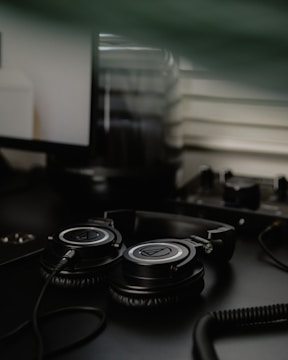 A pair of black over-ear headphones with a coiled cable resting on a dark desk. In the background, part of a computer monitor and some equipment can be seen, slightly out of focus, contributing to a workspace environment.