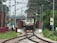 A train is approaching on a railway track surrounded by lush greenery. Electric poles and wires are prominently visible along the sides of the track. The scene appears to be in a rural or semi-rural area, with trees and shrubs adding to the natural environment.