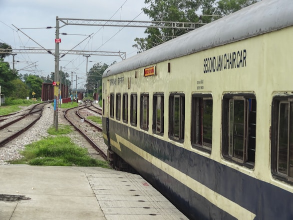 A passenger train travels along curved railway tracks surrounded by green vegetation and overhead power lines on a cloudy day. The side of the train is visible with windows and the words 'SECOND JAN CHAIR CAR' marked on it.