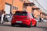 A red Volkswagen car is parked on a paved surface in front of a row of industrial buildings with orange brick walls and large garage doors. Several people are gathered around, some standing near other parked cars, while others appear to be walking away. The scene suggests a busy environment, possibly a car meet or event.