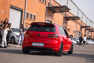 A red Volkswagen car is parked on a paved surface in front of a row of industrial buildings with orange brick walls and large garage doors. Several people are gathered around, some standing near other parked cars, while others appear to be walking away. The scene suggests a busy environment, possibly a car meet or event.