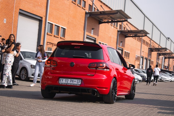 A red Volkswagen car is parked on a paved surface in front of a row of industrial buildings with orange brick walls and large garage doors. Several people are gathered around, some standing near other parked cars, while others appear to be walking away. The scene suggests a busy environment, possibly a car meet or event.