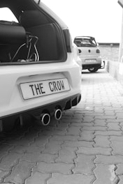 A close-up of the rear end of a white car with a modified exhaust and the license plate 'THE CROW'. Another car is parked further ahead in the background on a paved area.
