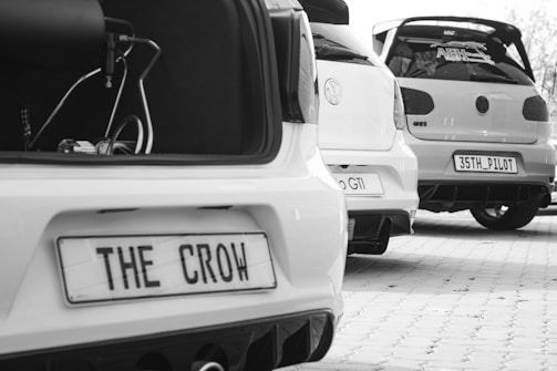 Three modified cars are parked in a line, capturing their rear ends. Each car has custom license plates and visible enhancements like spoilers. The scene is monochrome, highlighting the details of the vehicles.