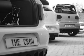 Three cars are parked in a row, with a focus on their rear ends. The nearest car's trunk is open, revealing part of its audio system. It has a custom license plate reading 'THE CROW'. The area is paved, and the cars appear to be showcased.