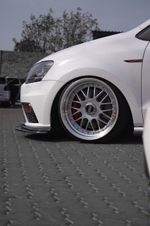 A close-up view of a white car with sporty alloy wheels parked on a paved surface. The wheel design is intricate, featuring multiple spokes, and the brake caliper is visible in red. The background includes a slightly blurred outdoor setting with trees and a building.