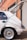 A white hatchback car with a sleek design and shiny chrome custom wheels is parked in a sunny industrial setting. The back door is open, revealing a spacious cargo area. The vehicle is positioned next to a brick wall and a closed metal garage door, suggesting an urban environment.