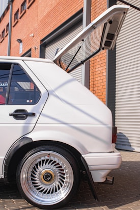 A white hatchback car with a sleek design and shiny chrome custom wheels is parked in a sunny industrial setting. The back door is open, revealing a spacious cargo area. The vehicle is positioned next to a brick wall and a closed metal garage door, suggesting an urban environment.