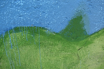 A close-up view of a textured wall that appears to be painted with two primary colors. The upper portion is painted blue, resembling a sky, and the lower portion is painted green, depicting a hilly landscape. There are visible drips of blue paint running down into the green area.