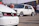 A classic white Volkswagen GTI car is parked on the pavement, showcasing its custom wheels and design. In the foreground, part of another Volkswagen is visible, displaying the GTI badge. Several people are standing and walking around in the background, suggesting a car show or gathering.