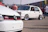 A classic white Volkswagen GTI car is parked on the pavement, showcasing its custom wheels and design. In the foreground, part of another Volkswagen is visible, displaying the GTI badge. Several people are standing and walking around in the background, suggesting a car show or gathering.