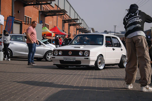 A white Volkswagen GTI car is parked on a paved surface, surrounded by people and various cars. Several individuals are interacting and observing the car, which is the focal point. There are brick buildings in the background and colorful tents, suggesting a car event or gathering.