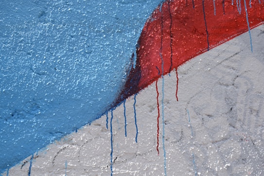 A textured wall features abstract art with vibrant colors. The upper section is painted blue with noticeable paint drips flowing down. Below the blue area, vivid red is layered over white, with the same dripping effects indicating a dynamic creation process.