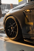 A sleek, modern car with a carbon fiber body finish is parked, partially captured from an angle that highlights its front side and large alloy wheels. The car has yellow detailing and is situated in a well-lit area, possibly a garage, with some background elements like another car and a person visible.