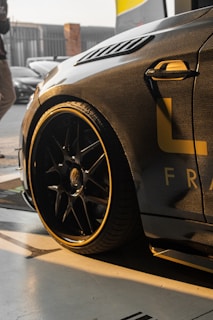 A sleek, modern car with a carbon fiber body finish is parked, partially captured from an angle that highlights its front side and large alloy wheels. The car has yellow detailing and is situated in a well-lit area, possibly a garage, with some background elements like another car and a person visible.