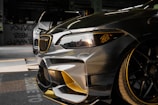 Close-up view of a customized car with a sleek design featuring carbon fiber and gold accents. In the background, another car is partially visible. The setting appears to be indoors with graffiti on the wall saying 'YOU'VE BEEN CLIMAX' and other details visible in the shadows.