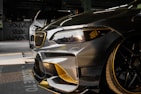 Close-up view of a customized car with a sleek design featuring carbon fiber and gold accents. In the background, another car is partially visible. The setting appears to be indoors with graffiti on the wall saying 'YOU'VE BEEN CLIMAX' and other details visible in the shadows.