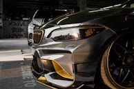 Close-up view of a customized car with a sleek design featuring carbon fiber and gold accents. In the background, another car is partially visible. The setting appears to be indoors with graffiti on the wall saying 'YOU'VE BEEN CLIMAX' and other details visible in the shadows.