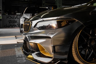 Close-up view of a customized car with a sleek design featuring carbon fiber and gold accents. In the background, another car is partially visible. The setting appears to be indoors with graffiti on the wall saying 'YOU'VE BEEN CLIMAX' and other details visible in the shadows.