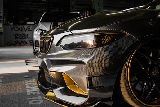 Close-up view of a customized car with a sleek design featuring carbon fiber and gold accents. In the background, another car is partially visible. The setting appears to be indoors with graffiti on the wall saying 'YOU'VE BEEN CLIMAX' and other details visible in the shadows.
