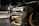 Close-up view of a customized car with a sleek design featuring carbon fiber and gold accents. In the background, another car is partially visible. The setting appears to be indoors with graffiti on the wall saying 'YOU'VE BEEN CLIMAX' and other details visible in the shadows.