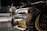 Close-up view of a customized car with a sleek design featuring carbon fiber and gold accents. In the background, another car is partially visible. The setting appears to be indoors with graffiti on the wall saying 'YOU'VE BEEN CLIMAX' and other details visible in the shadows.